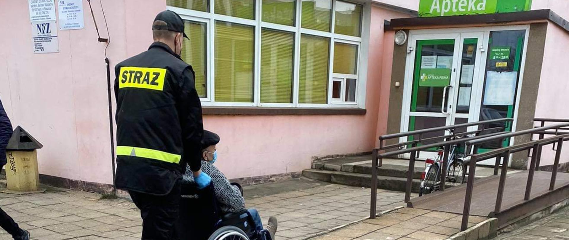 Na zdjęciu druh OSP transportujący na wózku inwalidzkim osobę starszą do punktu szczepień