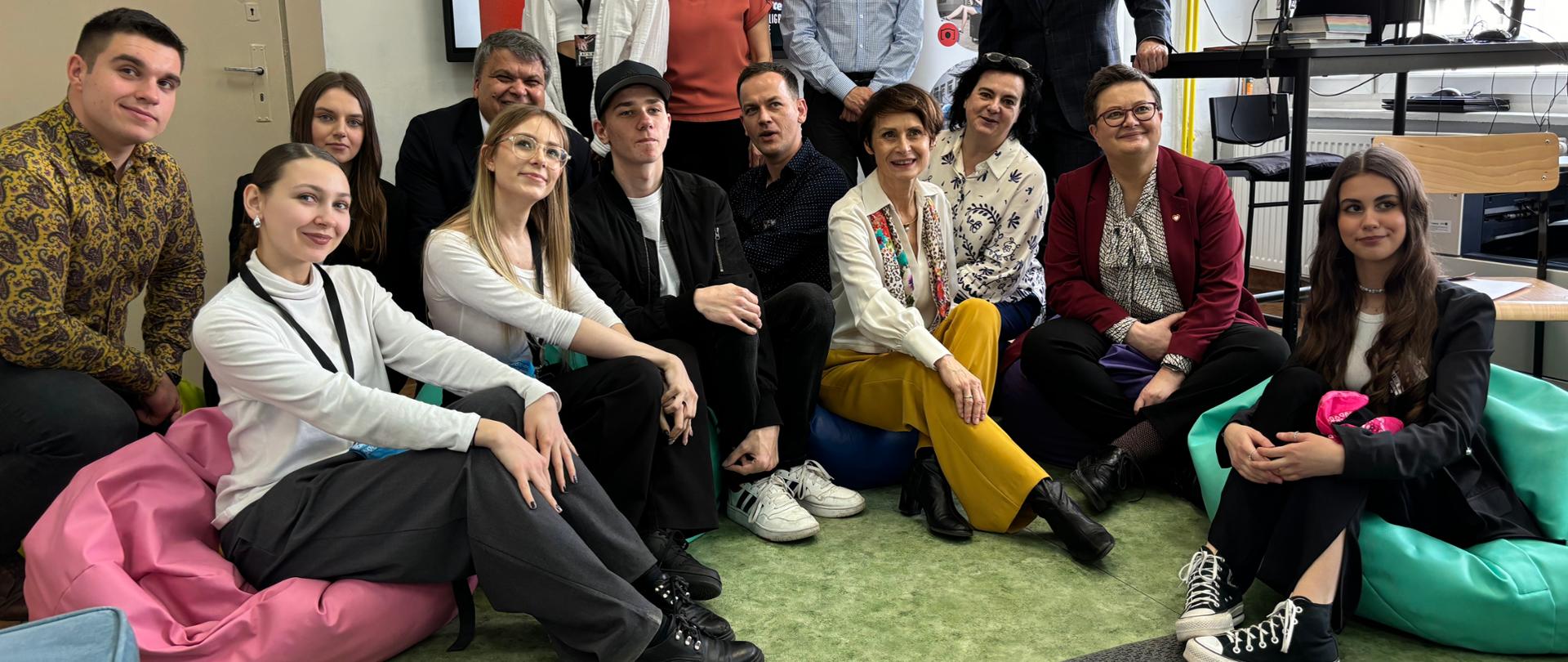 Wiceminister Lubnauer pozuje z uczniami i nauczycielami do zdjęcia w sali lekcyjnej Zespołu Szkół Ekonomicznych w Opolu. Część osób, wraz z wiceminister, siedzi na pufach położonych na ziemi, a pozostałe osoby są ustawione za ich plecami. 