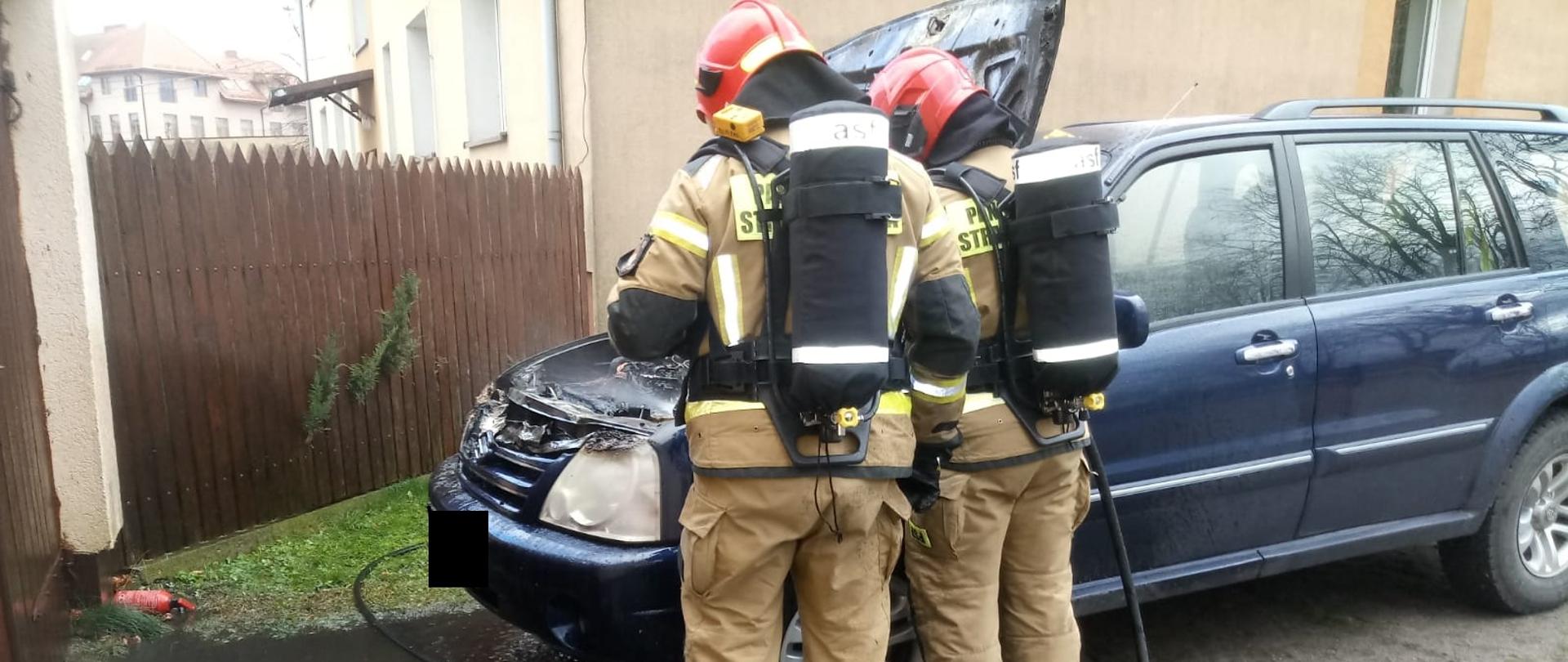 2 strażaków na podwórku przed garażem gasi palące się auto osobowe. 