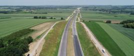 Fotografia zrobiona z lotu ptaka. Widoczne są dwa pasy nowobudowanej drogi pomiędzy polami uprawnymi – na całą długość zdjęcia, trwale od siebie oddzielone środkowym pasem zieleni – dla przeciwnych kierunków ruchu. Po dwóch stronach widoczne drogi serwisowe, rozciągające się na długości docelowych dróg. Malowniczy krajobraz – dużo wzniesień, dużo zieleni, drzew, piękna panorama.