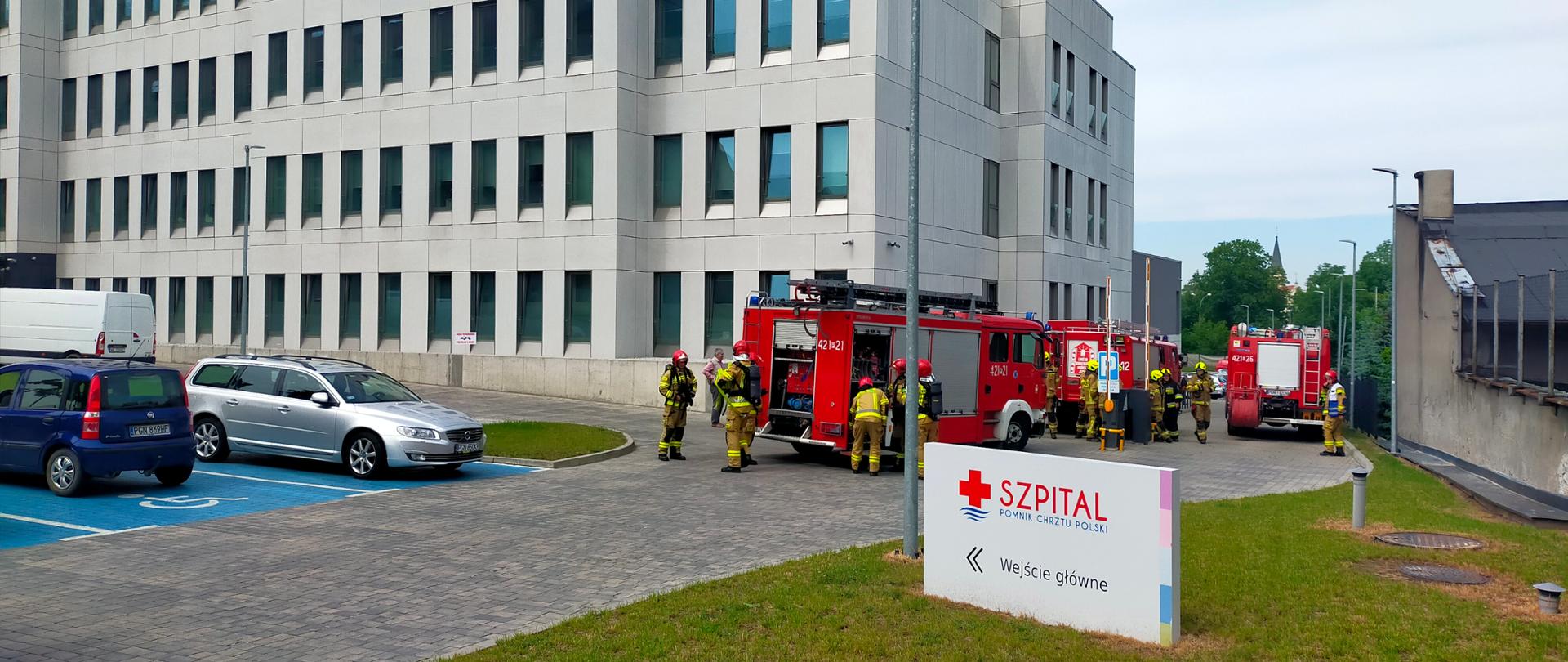 Na zdjęciu widać budynek szpitala przed który podjechały samochody pożarnicze. Strażacy zaczynają przygotowywać sprzęt do działań
