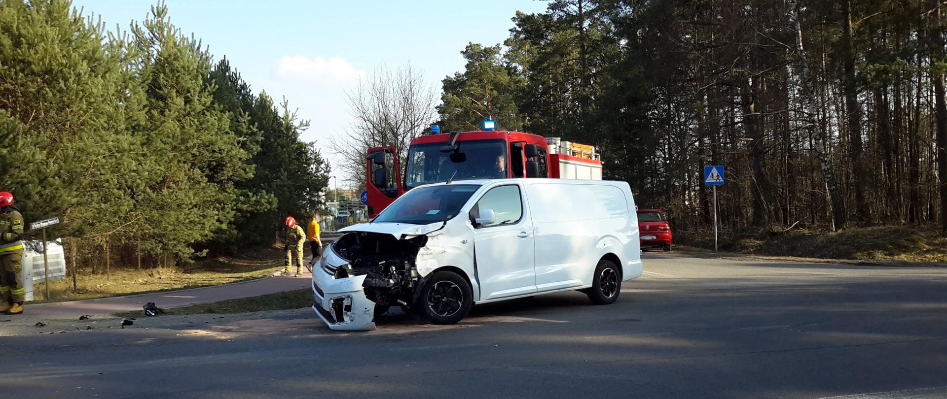 Samochód dostawczy z rozbitym przodem na drodze. Za nim wóz strażacki. Wokół strażacy prowadzą działania.