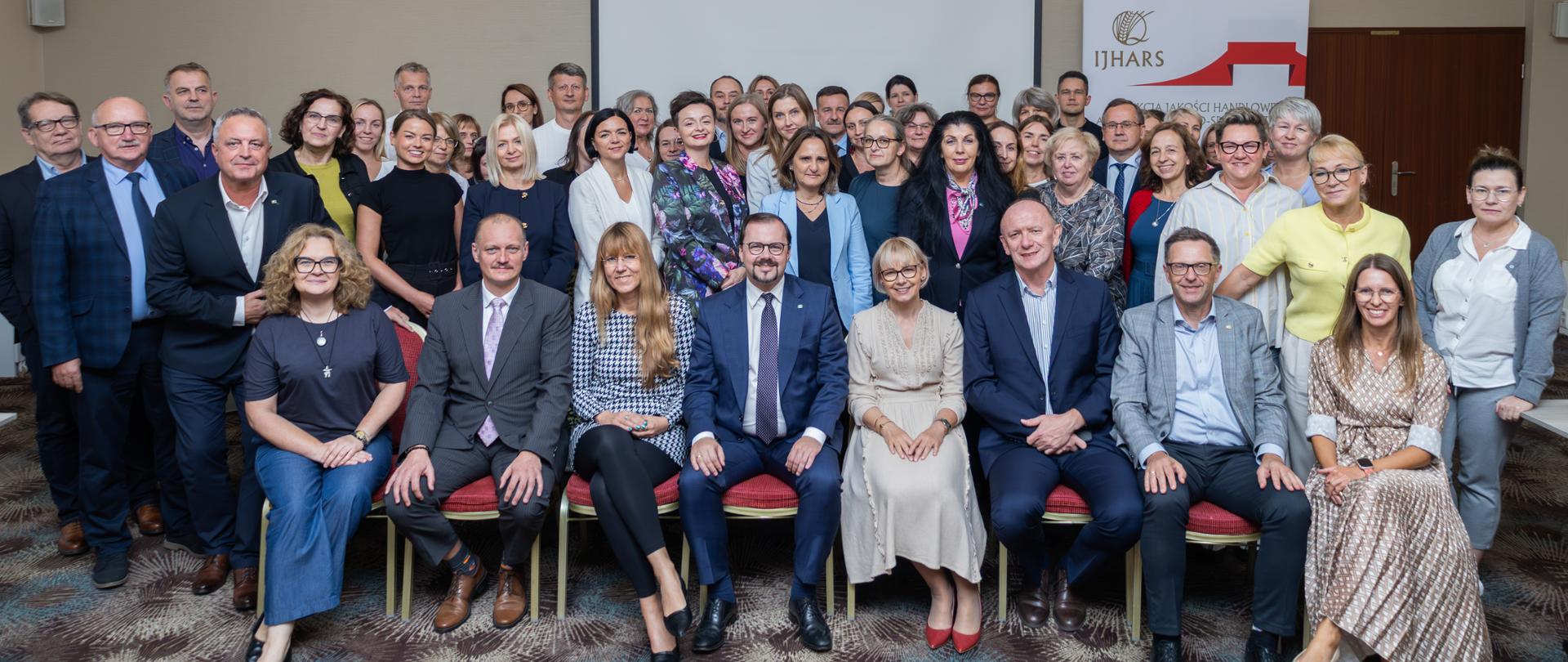 Ujęcie przedstawiające grupę ludzi, którzy są ubrani elegancko; tworzą kilka rzędów, rząd na pierwszym planie zajmuje miejsca siedzące. Wszyscy patrzą w obiektyw i delikatnie się uśmiechają.