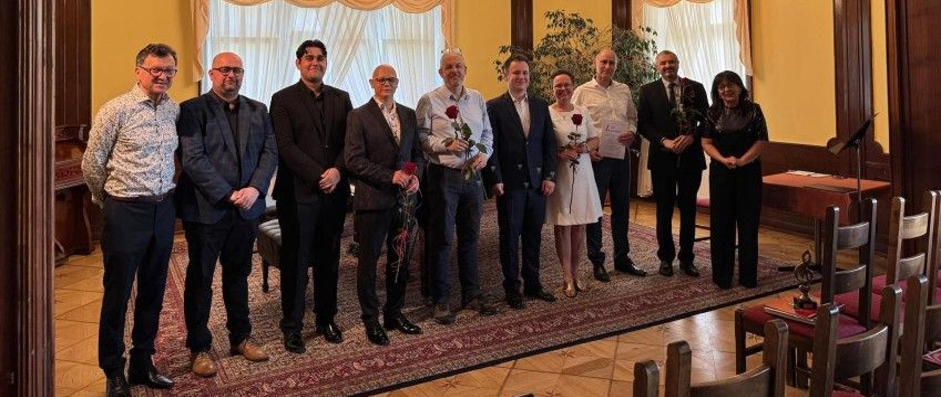 Zdjęcie kolorowe. W długim, reprezentacyjnym pomieszczeniu, z drewnianymi panelami i żółtymi ścianami, grupa dziesięciu osób, w tym dwie kobiety i ośmiu mężczyzn. Stoją oni na czerwonym dywanie z wzorem, na tle wysokich okien zasłoniętych jasnymi firankami. Większość mężczyzn ubrana jest w garnitury, a jedna kobieta w białą sukienkę, druga w ciemny strój. Niektóre osoby trzymają czerwone róże. Na ścianie za nimi godło Polski. Po prawej stronie zdjęcia widoczne są puste krzesła.
