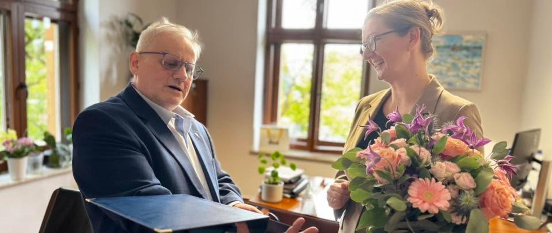 Mężczyzna w garniturze trzyma w ręku tabliczkę pamiątkową obok niego stoi kobieta z kwiatami w rękach 
