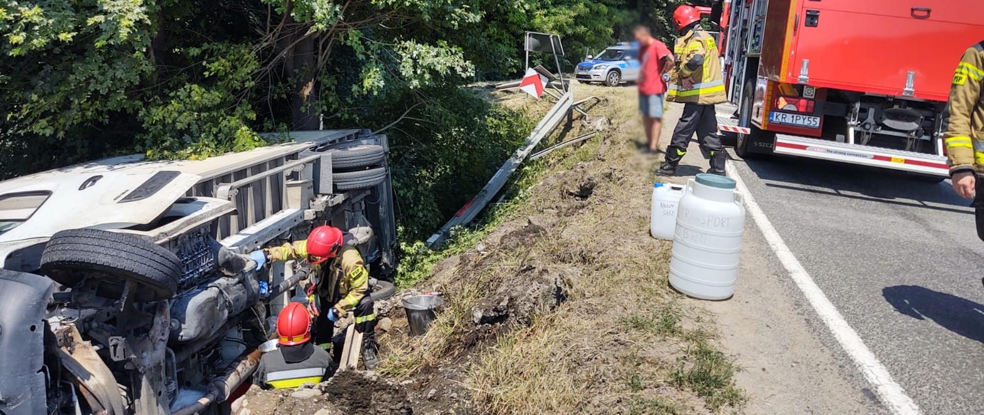 Samochód dostawczy poza drogą. Obok niego działania prowadzą strażacy. Na drodze samochód pożarniczy, beczka strażacy oraz osoba postronna.