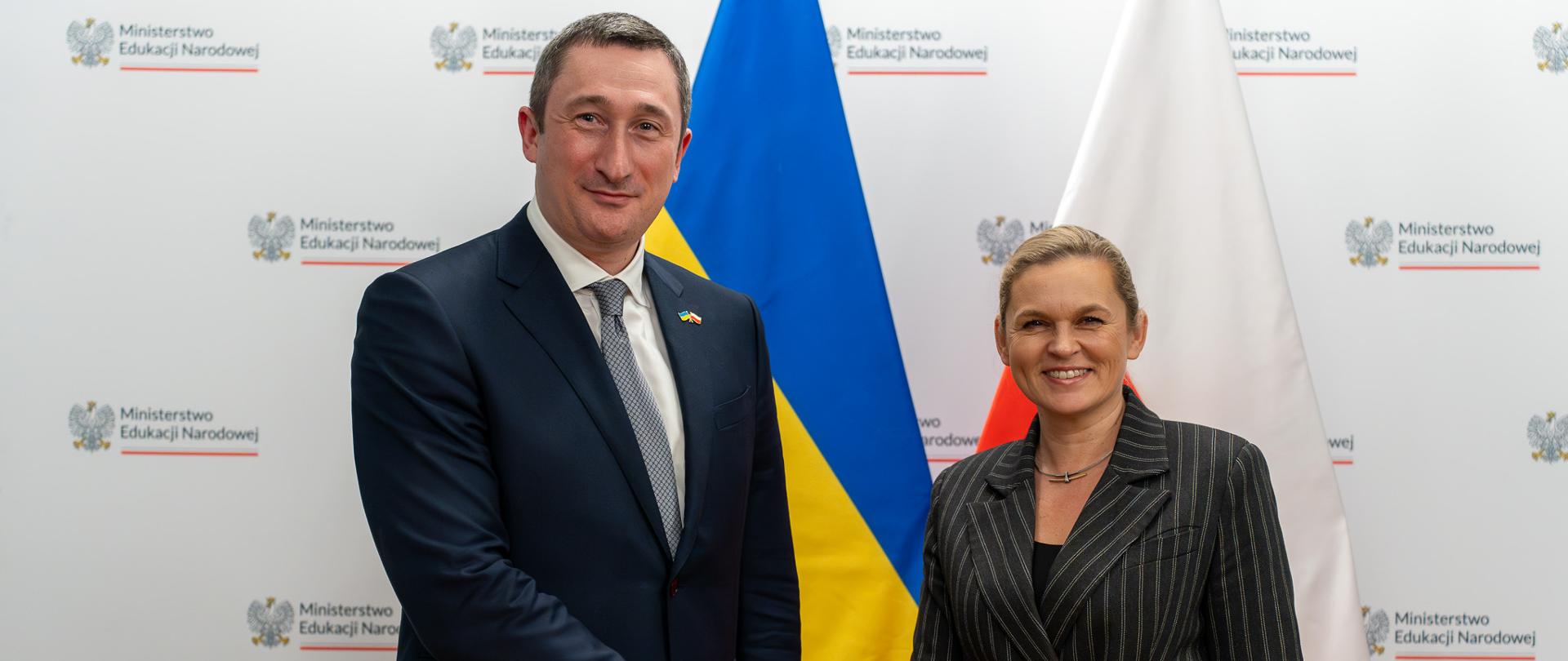 Na tle białej ścianki z napisem Ministerstwo Edukacji Narodowej oraz flag Ukrainy i Polski stoi po prawej minister Barbara Nowacka i podaje rękę mężczyźnie w garniturze wicepremierowi Ukrainy Ołeksijowi Czernyszowowi 