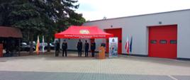 Widać strażaków pod czerwonym namiotem, jeden z nich przemawia przy mównicy, w tle budynek garażowy