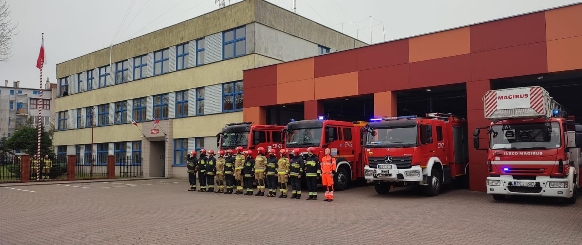 Zmiana służbowa na tle samochodów. Dowódca salutuje, na końcu pododdziału strażaków ratownik medyczny. Z lewej strony widać strażaków salutujących pod masztem.