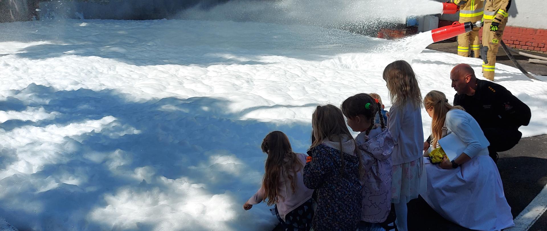 Dwóch strażaków ubranych w hełmy i ubrania specjalne trzyma w rękach wytwornice pianowe. Na ziemi rozlana jest piana ciężka, której przyglądają się dzieci.

