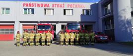 Zdjęcie przedstawia strażaków ustawionych w dwuszeregu przed pojazdami pożarniczymi, oddających hołd zmarłemu strażakowi. Przy samochodach włączone sygnały świetlno-dźwiękowe. W tle strażnica strażacka