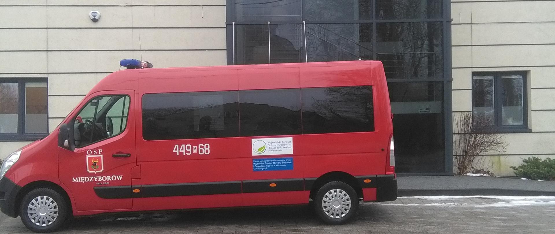 Na tle budynku przychodni wyznaczonej na punkt szczepień przeciwko COVID-19 stoi na parkingu samochód typu bus koloru czerwonego przeznaczony do przewozu osób należący do OSP Międzyborów