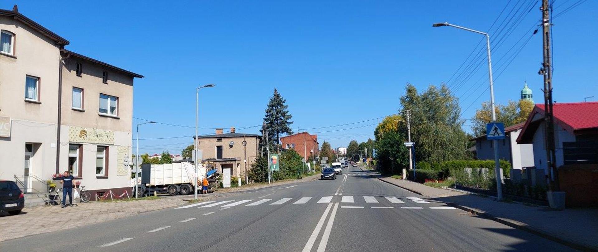 Skrzyżowanie DK11 z ul. Główną w Tarnowskich Górach. Po lewej stronie budynek z punktami o charakterze usługowo-handlowym oraz mieszkalnym. Patrząc z tej perspektywy jadąc na wprost dojedziemy do skrzyżowania z DK78 i ul. Bytomską. Niebo jest błękitne i bezchmurne, na fotografii ukazana jest pora letnia.