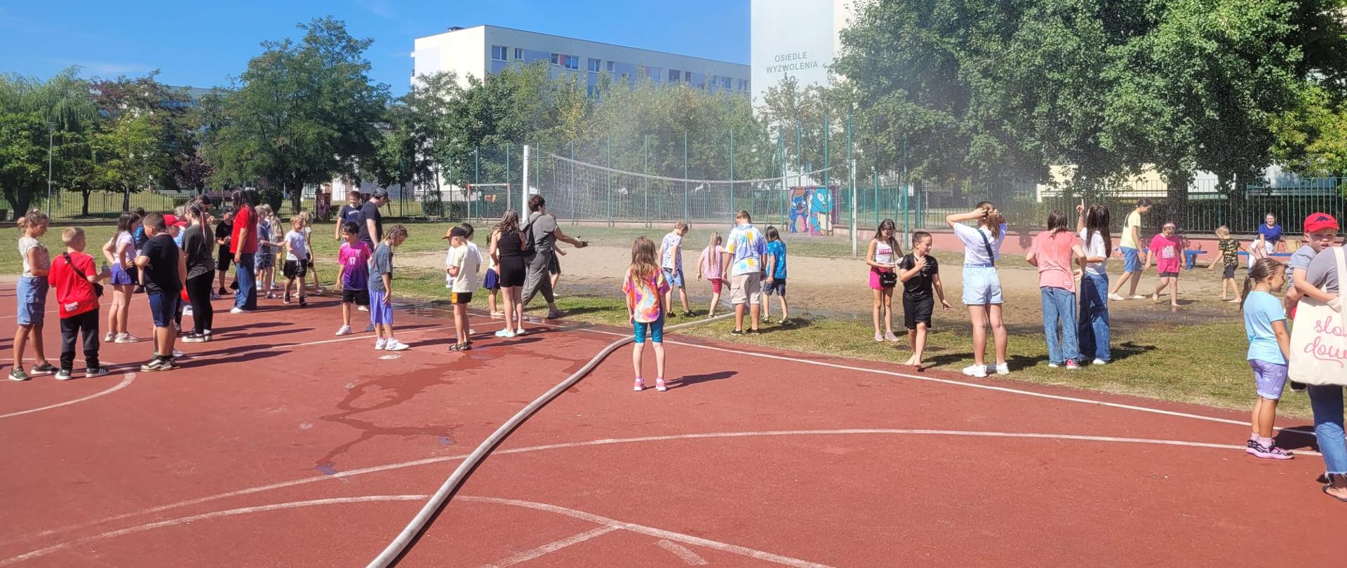 Dzieci i dorośli bawią się na tartanowym boisku oraz piaszczystym boisku do siatkówki. W centralnym punkcie rozłożona jest kurtyna wodna, z której leci woda. 