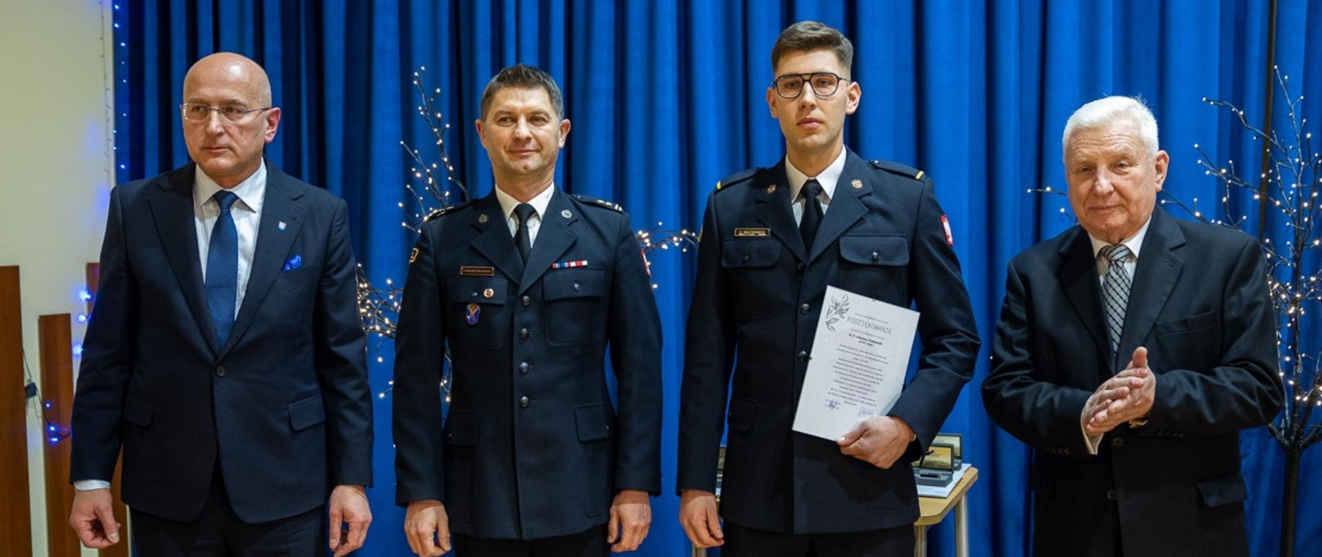 dwóch strażaków stoi obok nich stoją inne dwie osoby za nimi niebieskie tło