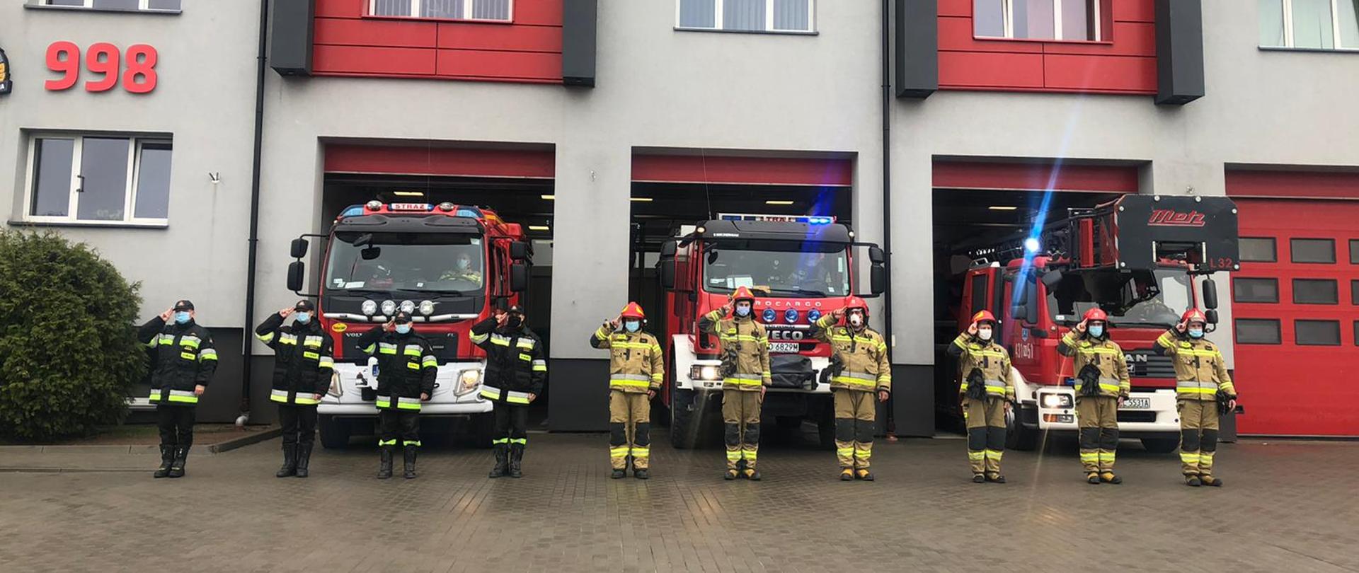 Na zdjęciu funkcjonariusze Straży Pożarnej oddający honor na tle budynku Komendy Powiatowej Państwowej Straży Pożarnej w Ełku i pojazdów pożarniczych.