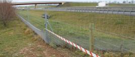 Na zdjęciu widać zamontowane ogrodzenie z siatki na drewnianych kołkach, na siatce na części ogrodzenie taśma biało-czerwona. Za ogrodzeniem widać rów przydrożny, za nim drogę, a w oddali wiadukt nad drogą.