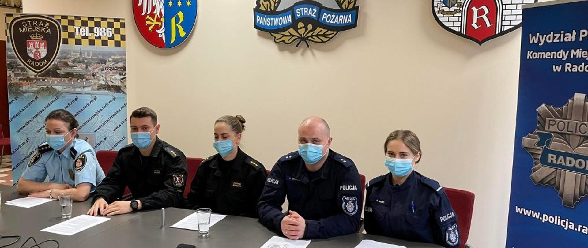 Zdjęcie wykonane w sali konferencyjnej w obiekcie Komendy Miejskiej Państwowej Straży Pożarnej w Radomiu. Przy stole siedzą trzy kobiety oraz dwóch mężczyzn. Są oni funkcjonariuszami Straży Miejskiej w Radomiu, Komendy Miejskiej Państwowej Straży Pożarnej w Radomiu oraz Komendy Miejskiej Policji w Radomiu. Nad nimi na ścianie znajdują się herb miasta Radomia oraz logo Państwowej Straży Pożarnej. Po ich lewej stronie znajduje się dodatkowo baner z logiem Straży Miejskiej w Radomiu. Podobny baner zawierający logo Komendy Miejskiej Policji w Radomiu znajduje się na prawo od nich. 
