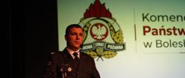 Funkcjonariusz Państwowej Straży Pożarnej w mundurze wyjściowym przemawia przy mównicy na tle prezentacji Komendy Powiatowej PSP w Bolesławcu.