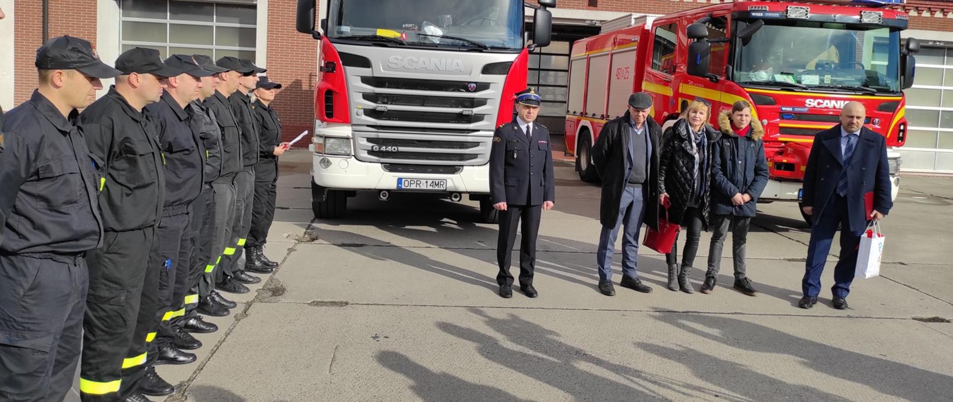 Zdjęcie przedstawia strażaków i osoby cywilne na uroczystym apelu na placu Komendy Powiatowej PSP. W tle samochody pożarnicze i budynek strażnicy.