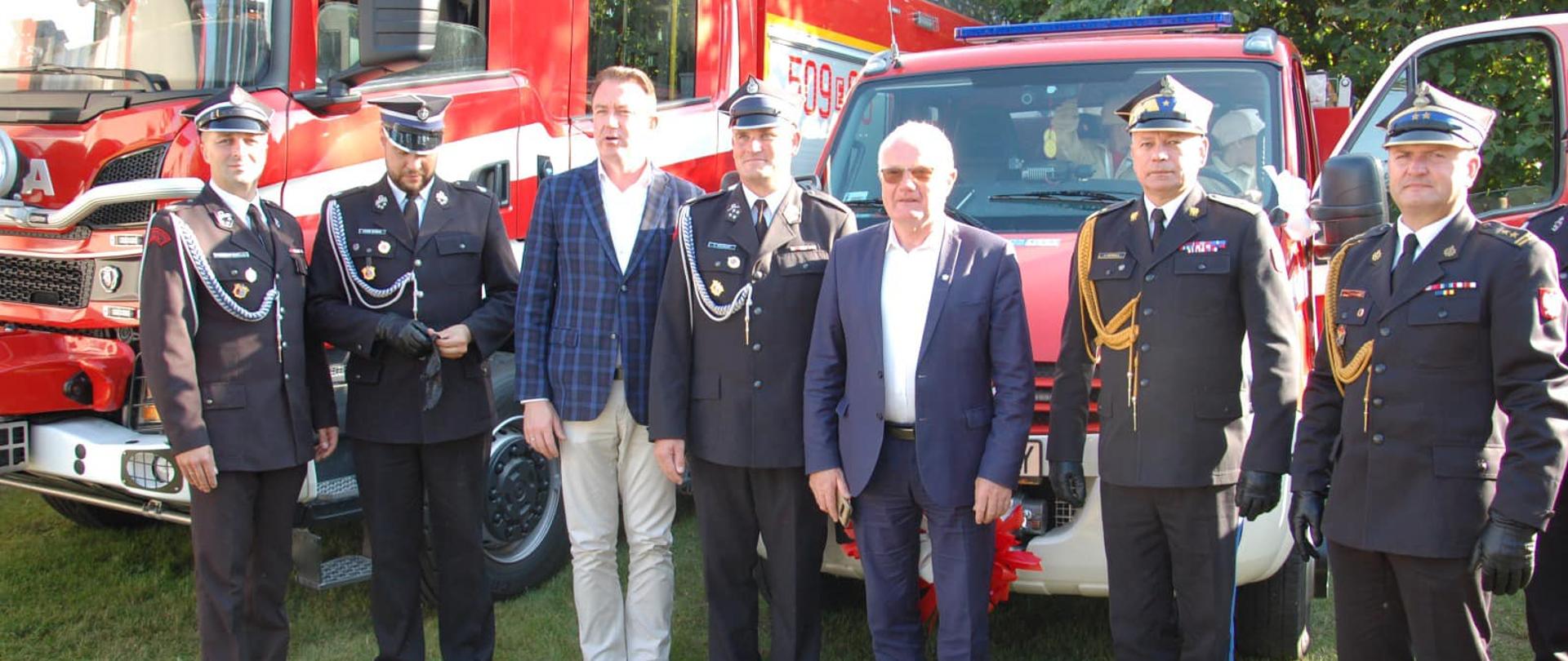 Zastępca Komendanta Głównego PSP - nadbrygadier Arkadiusz Przybyła pozuje do zdjęcia grupowego na tle pojazdów pożarniczych. Obok niego stoją druhowie OSP i samorządowcy.