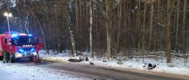 Na drodze w lesie stoi samochód strażacki przed nim lez torba medyczna koloru czerwonego na poboczy drogi uszkodzony motorower oraz porozbijane elementy po wypadku
