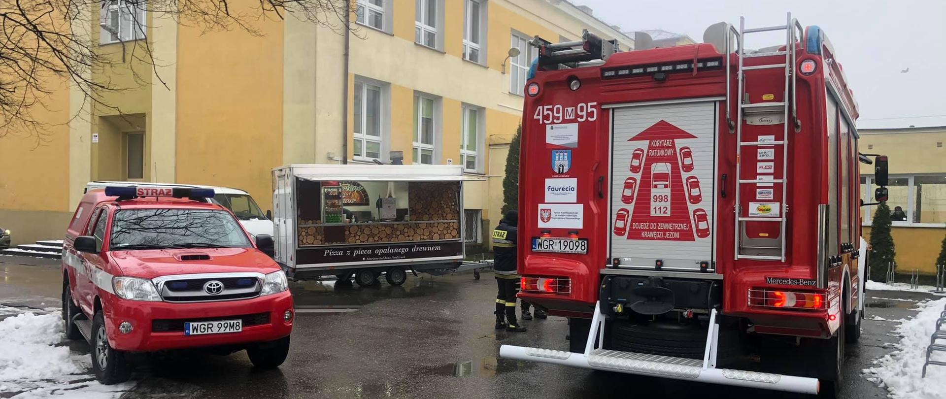 Zdjęcie przedstawia dwa samochody pożarnicze ustawione przed budynkiem szkoły w której to organizowana była zbiórka krwi. Na zdjęciu znajdują się pojazdy takie jak SLRR Toyota Hilux z KP PSP Grójec oraz GBA Mercedes Atego z OSP Grójec 