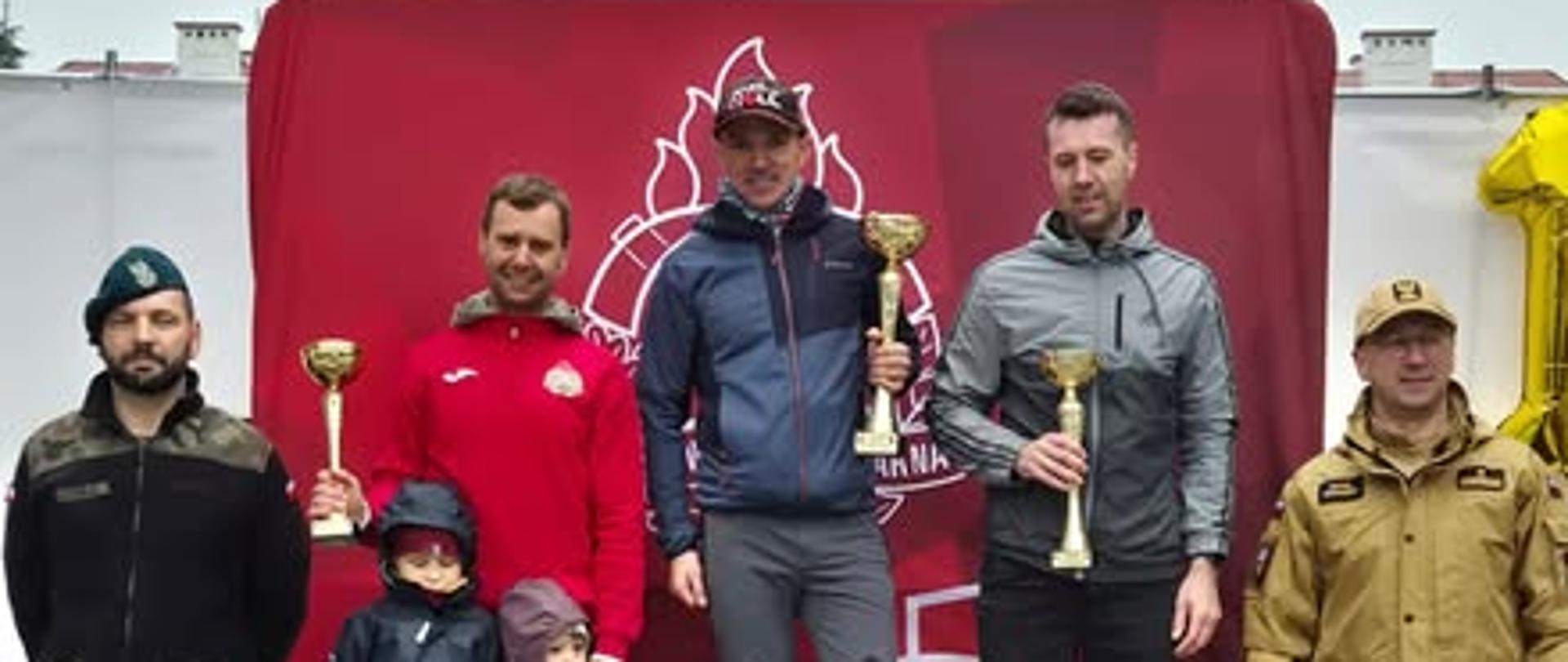 Fotografia przedstawia zwycięzców wyścigu. Stoją oni na podium, podium ma numery 1,2 i 3 odpowiadające miejscom zajętym przez zawodników. Jednemu zawodnikowi towarzyszą dzieci na podium. Z lewej strony stoi funkcjonariusz Wojska Polskiego, po prawej natomiast widoczny jest funkcjonariusz Państwowej Straży Pożarnej. Zwycięzcy z rękach trzymają pamiątkowe puchary. W tle widać logo PSP na czerwonym tle. 