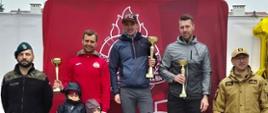 Fotografia przedstawia zwycięzców wyścigu. Stoją oni na podium, podium ma numery 1,2 i 3 odpowiadające miejscom zajętym przez zawodników. Jednemu zawodnikowi towarzyszą dzieci na podium. Z lewej strony stoi funkcjonariusz Wojska Polskiego, po prawej natomiast widoczny jest funkcjonariusz Państwowej Straży Pożarnej. Zwycięzcy z rękach trzymają pamiątkowe puchary. W tle widać logo PSP na czerwonym tle. 