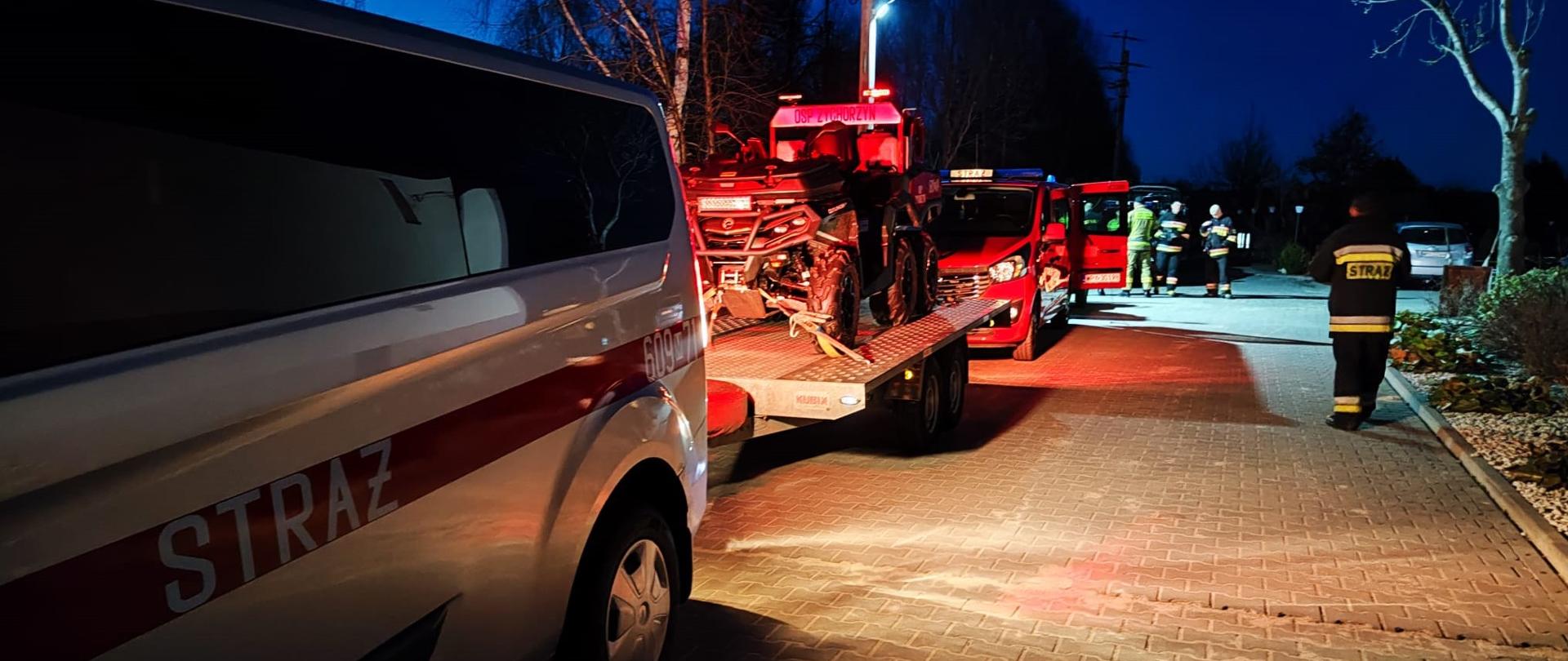 Zdjęcie przedstawia scenę nocną z pojazdami służb ratunkowych na utwardzonym placu lub drodze. Na pierwszym planie po lewej stronie znajduje się bok białego vana z czerwonym pasem i napisem "STRAŻ". Za nim, nieco dalej, widać czerwoną przyczepę z załadowanym na nią quadem. W tle, po prawej stronie zdjęcia, widoczna jest grupa strażaków w odblaskowych ubraniach i hełmach. Jeden ze strażaków idzie w kierunku centrum kadru, a na jego plecach widnieje napis "STRAŻ". Teren jest oświetlony światłami pojazdów i lamp ulicznych, co tworzy długie cienie i rzuca czerwone refleksy na ziemię. Po obu stronach drogi rosną drzewa, a w oddali widać ciemne budynki. Scena sugeruje interwencję lub przygotowanie do akcji ratowniczej prowadzonej przez straż pożarną.