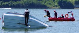Po prawej stronie wywrócona żaglówka z załogantem stawiającym ją na wodę. Po prawej stronie - ratownik z PSP w trakcie skoku do wody z łodzi ratowniczej.