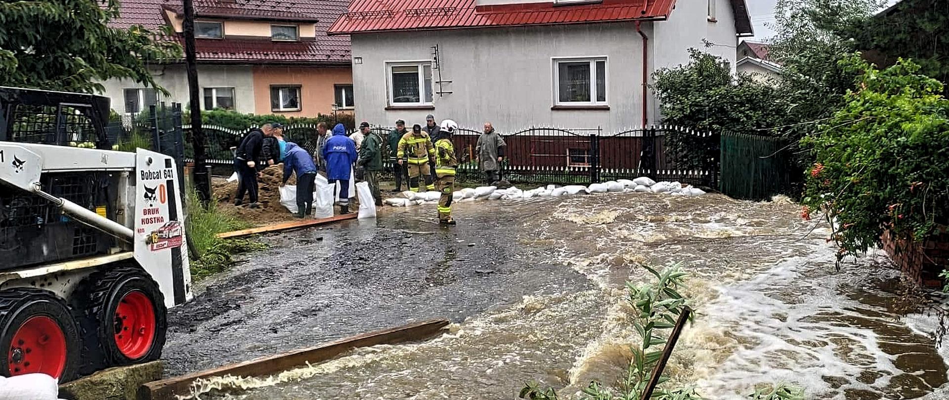 Na pierwszym planie mini koparka na drugim planie grupa jedenastu mężczyzn w tym dwóch strażaków w trakcie napełniania worków piaskiem. W tle budynki mieszkalne wzdłuż ulicy. W głównym kadrze rwący strumień wody płynący ulicą. 