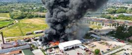 Widok z lotu ptaka na duży pożar w kompleksie przemysłowym. Z hali i placu składowego unoszą się gęste, czarne kłęby dymu oraz widać płomienie. Na miejscu jest wiele wozów strażackich, a strażacy prowadzą akcję gaśniczą. W tle rozciągają się tory kolejowe, pola i zabudowania.