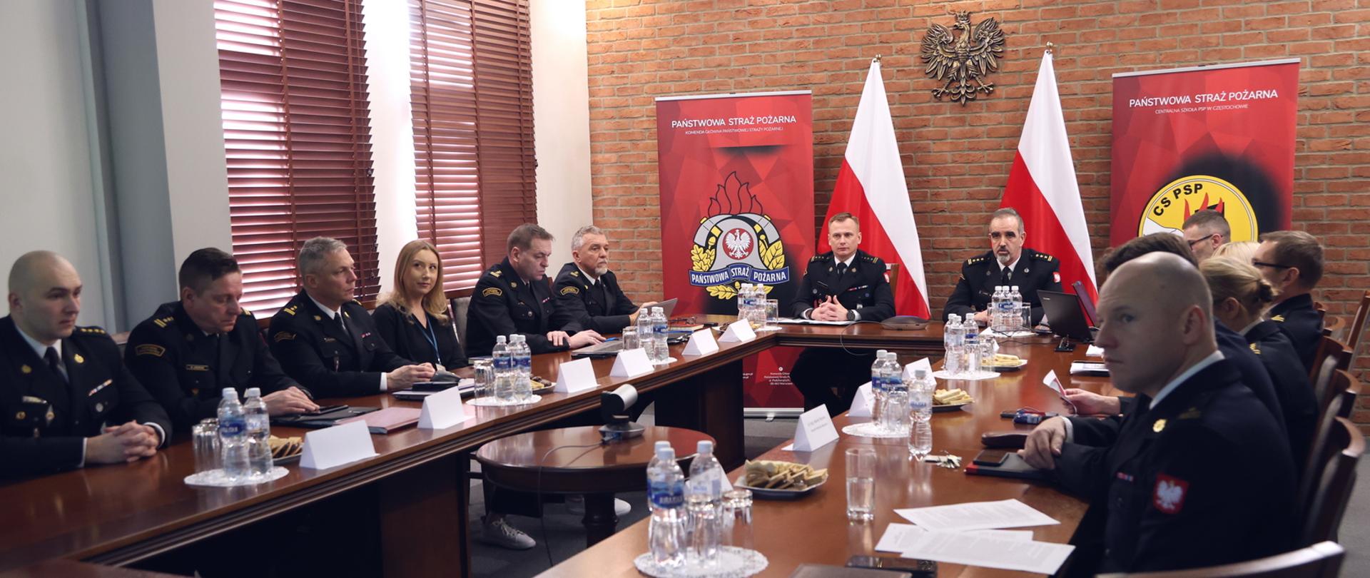 W sali konferencyjnej przy stołach ustawionych w formie prostokąta siedzi podczas narady p.o. Komendant Centralnej Szkoły PSP, funkcjonariusze kadry kierowniczej CS PSP oraz kobieta. W głębi stojące na stojakach dwie biało-czerwone flagi obok których widnieją banery z logo PSP oraz CS PSP. Na ścianie wiszący orzeł w koronie