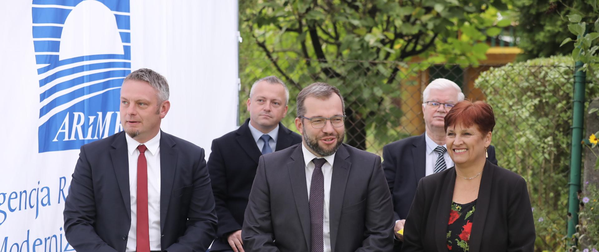 Trzy osoby siedzą na krzesłach. W środku minister Grzegorz Puda. W drugim rzędzie siedzą dwie inne osoby.