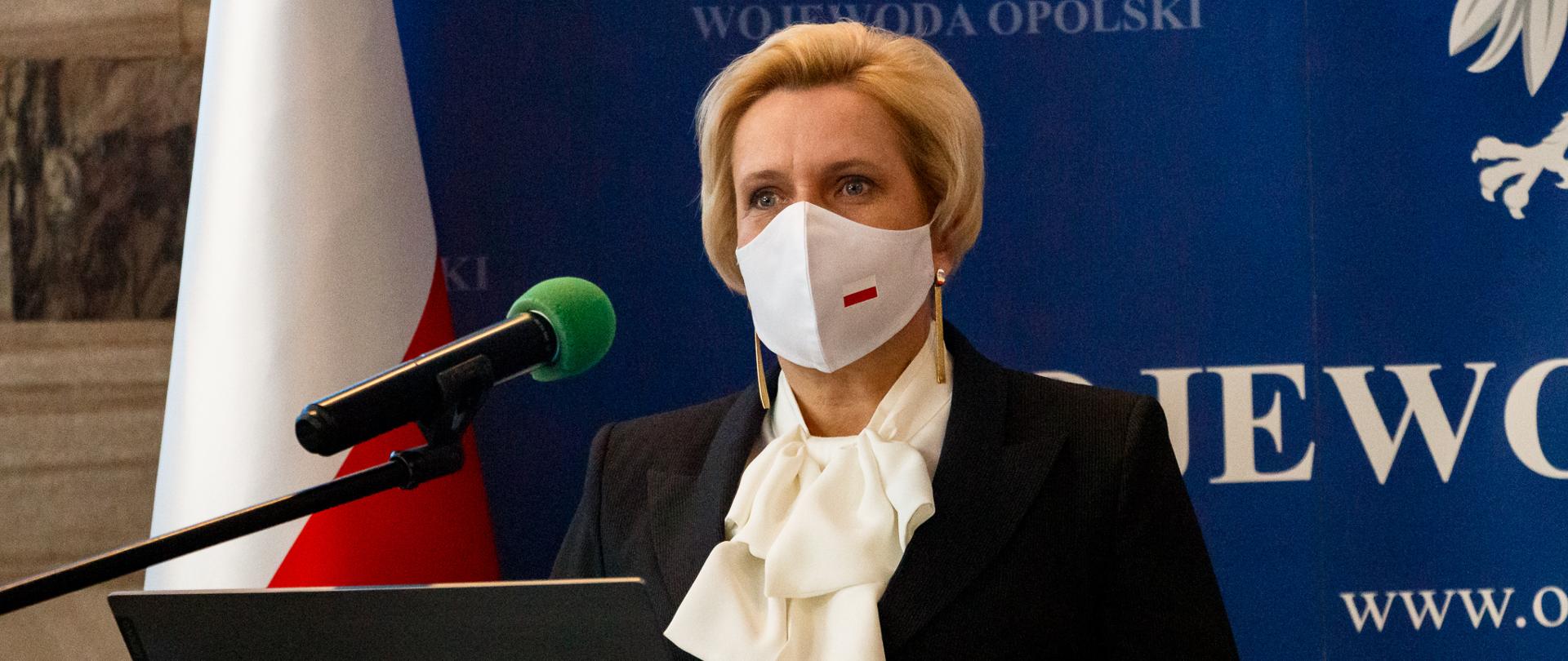 Na zdjęciu Wicewojewoda opolski Teresa Barańska na tle granatowej ścianki z napisem Wojewoda Opolski. W dali po jej prawej stronie widać stojącą biało-czerwoną flagę. Na pierwszym planie szklana mównica, na której stoi laptop. Przed nim stoi wicewojewoda opolski, która w dłoni trzyma pilot do laptopa. Na jej twarzy widać białą maseczkę ochronną z biało-czerwoną flagą. 