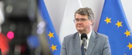 Mężczyzna w garniturze stoi przy mikrofonie za nimi flagi Unii Europejskiej.