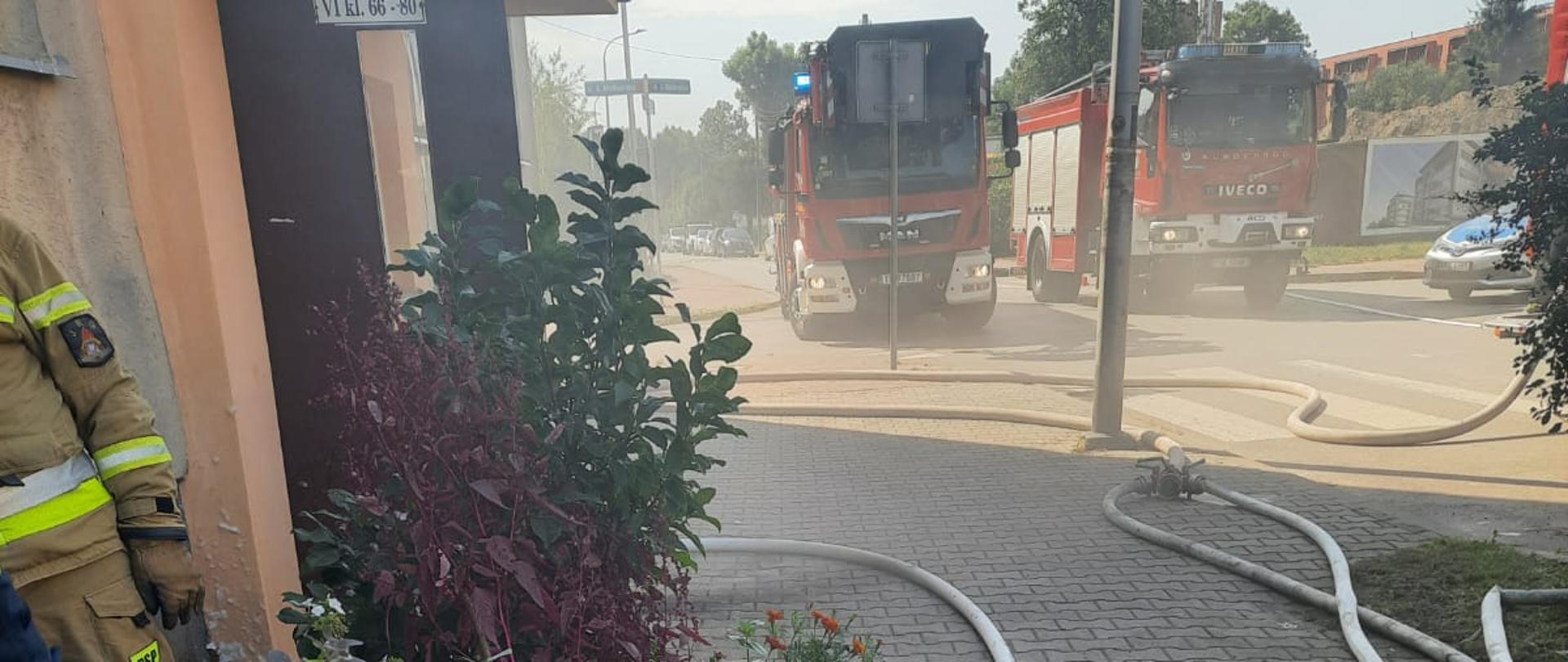 Zdjęcie przestawia dym wydobywający się z budynku wielorodzinnego. W tle stoją dwa samochody pożarnicze i radiowóz. Na ziemi przed wejściem leżą linie gaśnicze, a po lewej stronie przy ścianie mieszkania stoi strażak i jest wyrzucona duża ilość spalonych rzeczy.