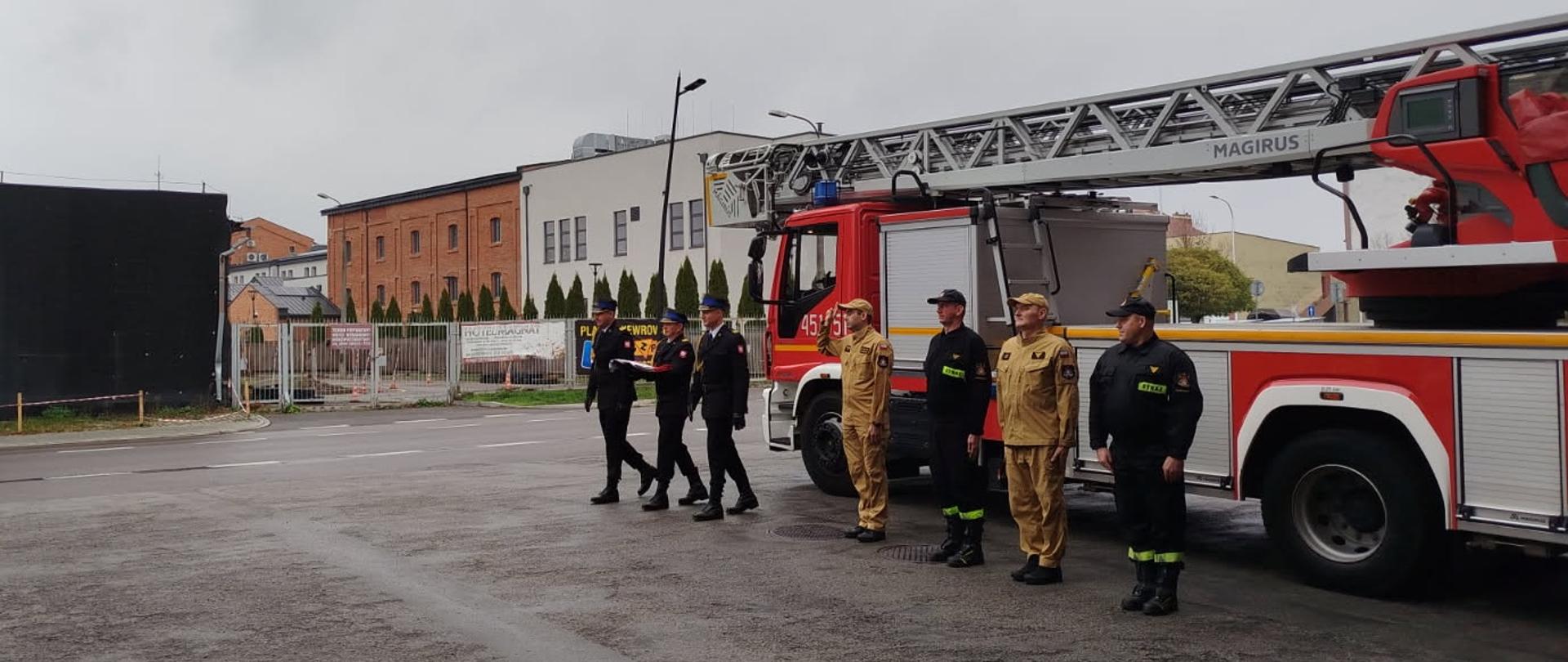 Poczet sztandarowy maszeruje by poidnieść flagę państwową. W tle samochód drabina pożarnicza.