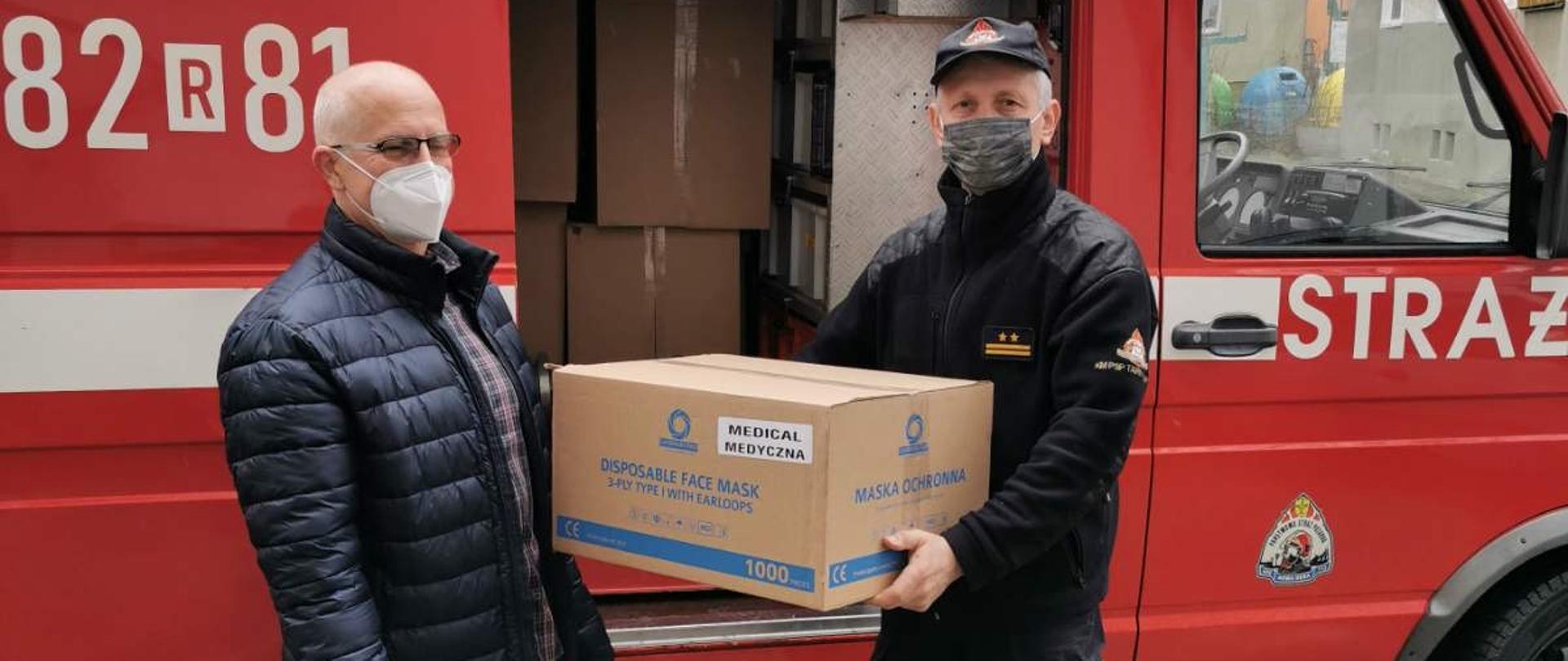 Dwóch mężczyzn stoi przed samochodem pożarniczym. Jeden z mężczyzn, który jest strażakiem, trzyma w rękach pudło kartonowe z napisem "maseczki ochronne".