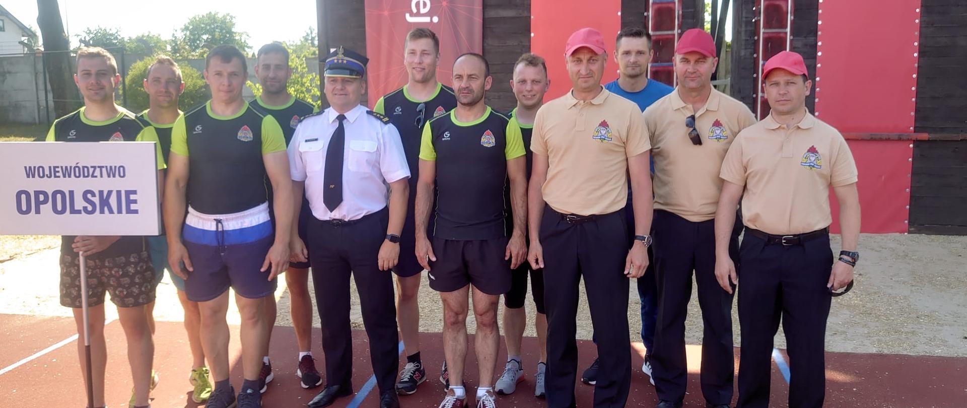 Wspólne zdjęcie zawodników z Opolszczyzny z Opolskim Komendantem Wojewódzkim PSP w Opolu st. bryg. Krzysztofem Kędrykiem oraz sędziami oddelegowanymi z KP PSP z terenu województwa opolskiego. Zdjęcie na tartanie tuż przy wbiegu na wspinalnię do hakówki. Zawodnicy w strojach sportowych. Sędziowie w jasnych koszulkach polo i ciemnych spodniach oraz z czerwonymi czapkami z daszkiem na głowach. Opolski Komendant Wojewódzki PSP w Opolu w mundurze wyjściowym z koszulą letnią.