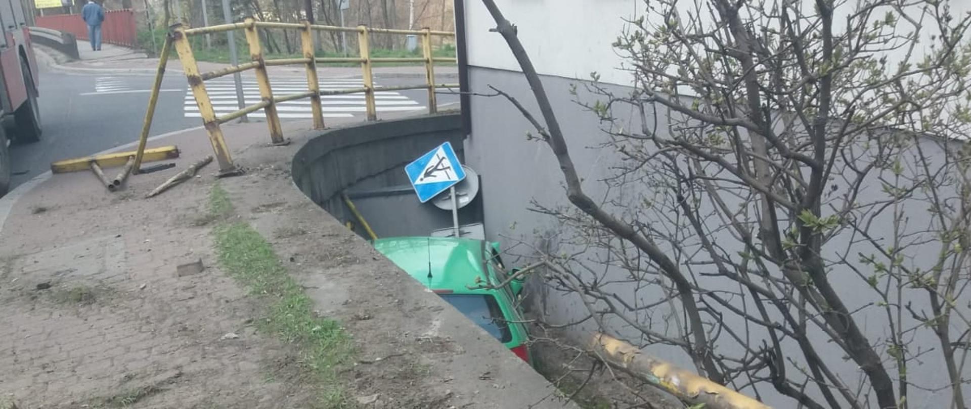 Zdjęcie przedstawia zielony samochód który wpadł pomiędzy mur oporowy budynku a jezdnie na głębokość ok 2 metrów