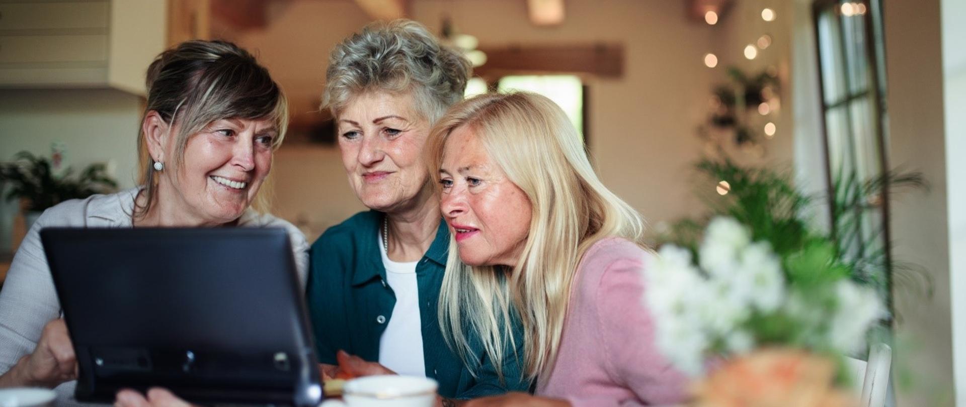 Trzy uśmiechnięte seniorki korzystają z tableta. 