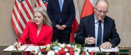 Deputy Minister of Climate and Environment Krzysztof Galos and Under Secretary of State Allison Hooker sign documents at a table during an official ceremony; both are dressed in formal attire—the woman in a red suit and the man in a business suit—while a third man in a suit stands behind them, with Polish and U.S. flags in the background.