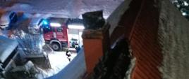 Zdjęcie przedstawia dach budynku, który w szczytowej części (kalenica) zniszczony jest przez pożar. Na dachu zalegają duże ilości śniegu. W samochód pożarniczy, który ma włączone niebieskie światła błyskowe.