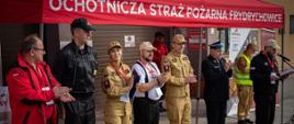 Widok z przodu. Przemówienie, współorganizatorzy – przedstawiciel Maltańskiej Służby Medycznej oraz przedstawiciele PSP. W tle namiot ochotniczej Straży Pożarnej we Frydrychowicach.
