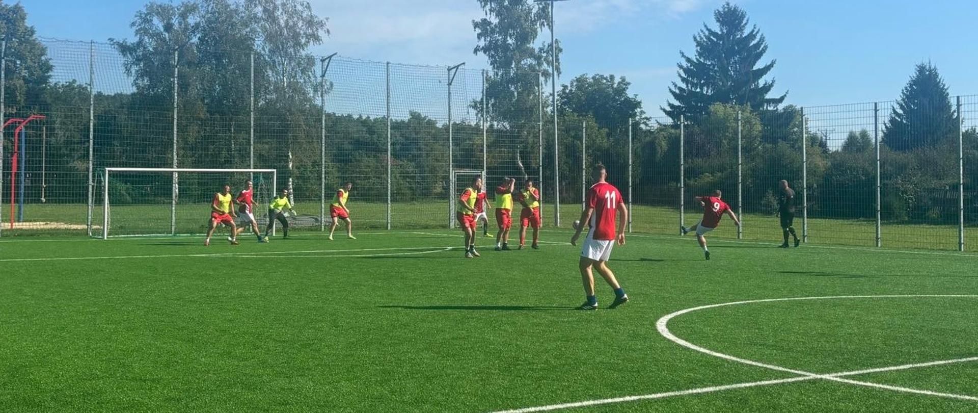 Mecz piłki nożnej strażaków na boisku Miejskiego Ośrodka Sportu i Rekreacji w Mławie. Zawodnicy w czerwonych i czarnych strojach rywalizują o piłkę w słoneczny dzień.