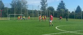 Mecz piłki nożnej strażaków na boisku Miejskiego Ośrodka Sportu i Rekreacji w Mławie. Zawodnicy w czerwonych i czarnych strojach rywalizują o piłkę w słoneczny dzień.