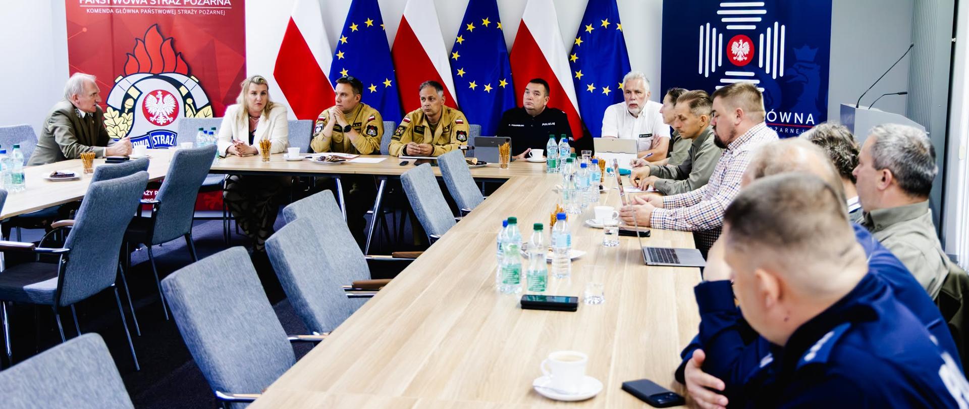 Widok ogólny sali konferencyjnej podczas spotkania przedstawicieli Państwowej Straży Pożarnej, Policji, Lasów Państwowych i innych instytucji. Uczestnicy siedzą przy długim stole. W tle znajdują się flagi Polski i Unii Europejskiej oraz banery Państwowej Straży Pożarnej.