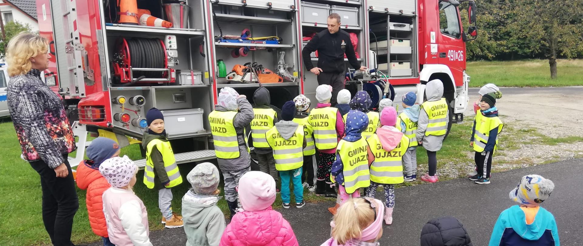 Na zdjęciu są dzieci, przedszkolanka i strażak na tle samochodu strażackiego